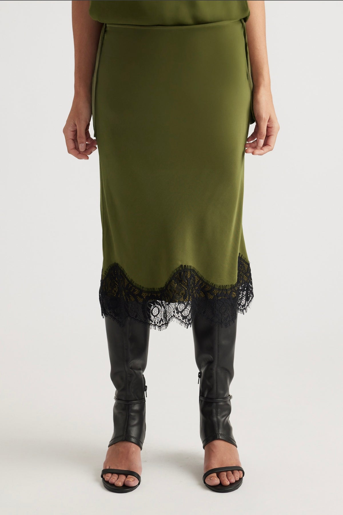 Front view of an olive green skirt with black lace trim paired with tall black leather heeled boots and sandals