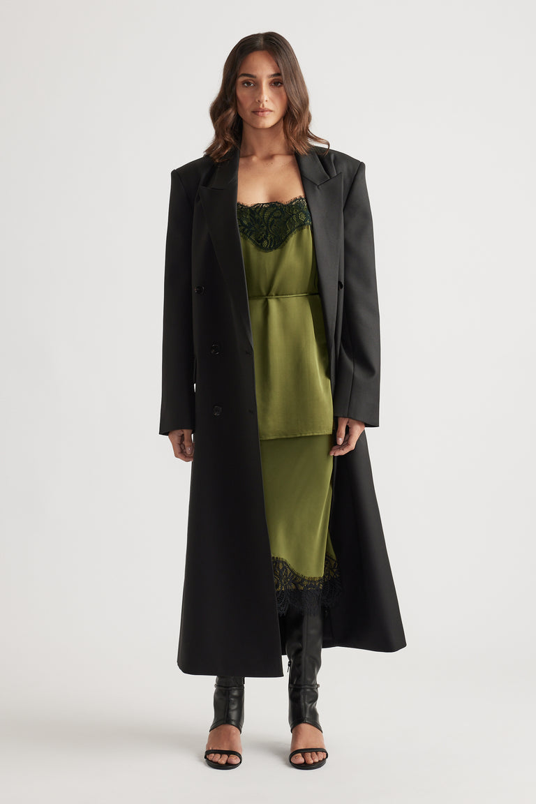 Long black tailored coat layered over a silky olive green lace-trimmed slip dress paired with open-toe black boots
