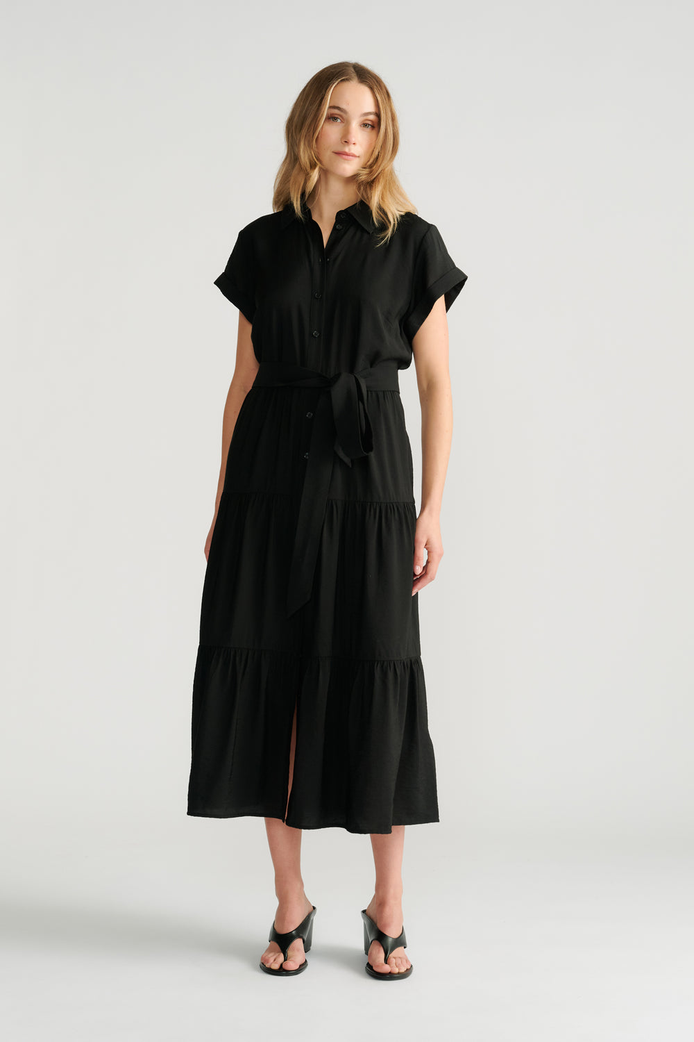Woman wearing a black midi shirt dress with rolled sleeves and a tied waist belt, paired with black heeled sandals.
