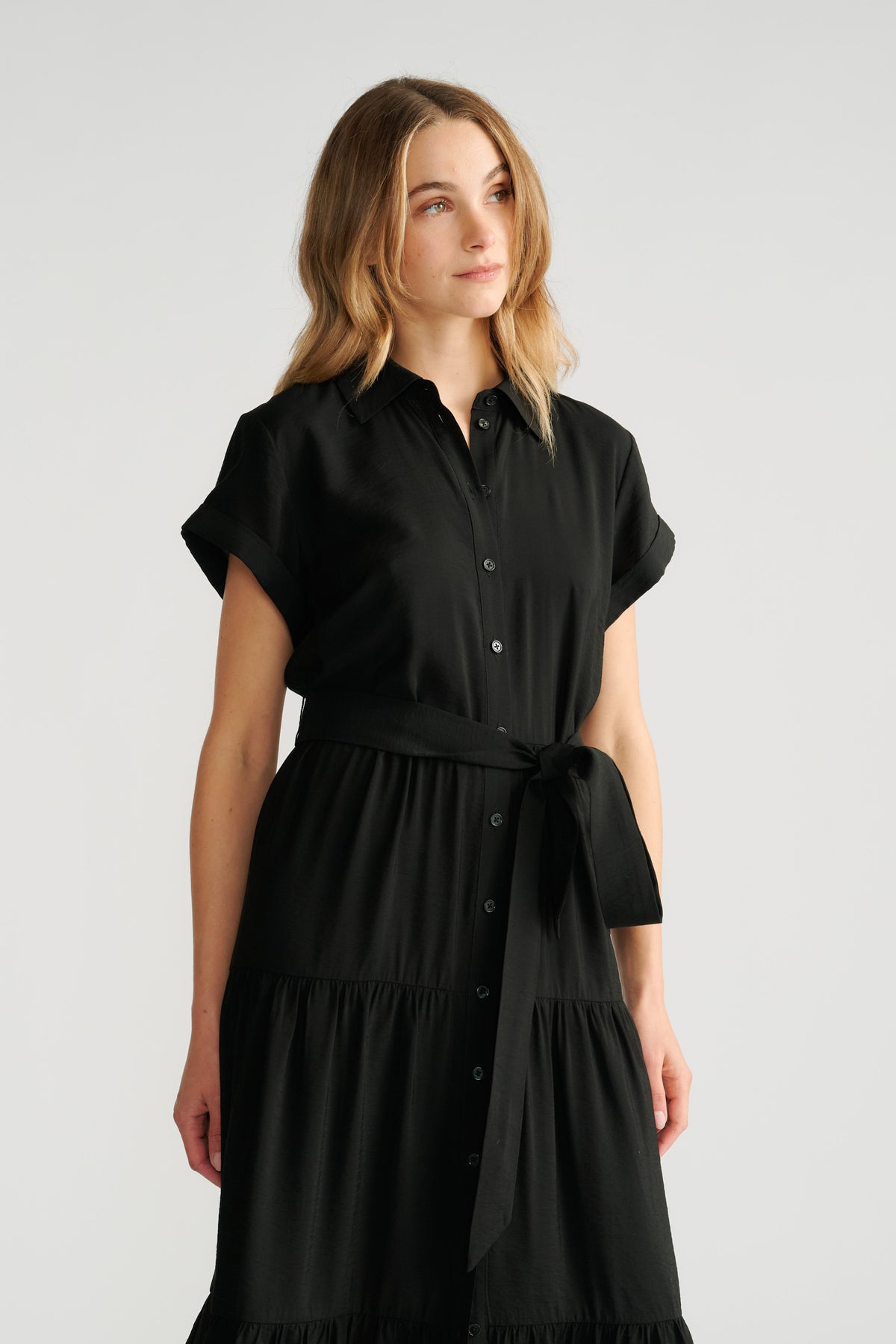 Woman wearing a black midi shirt dress with rolled sleeves and a tied waist belt, looking thoughtfully to the side.