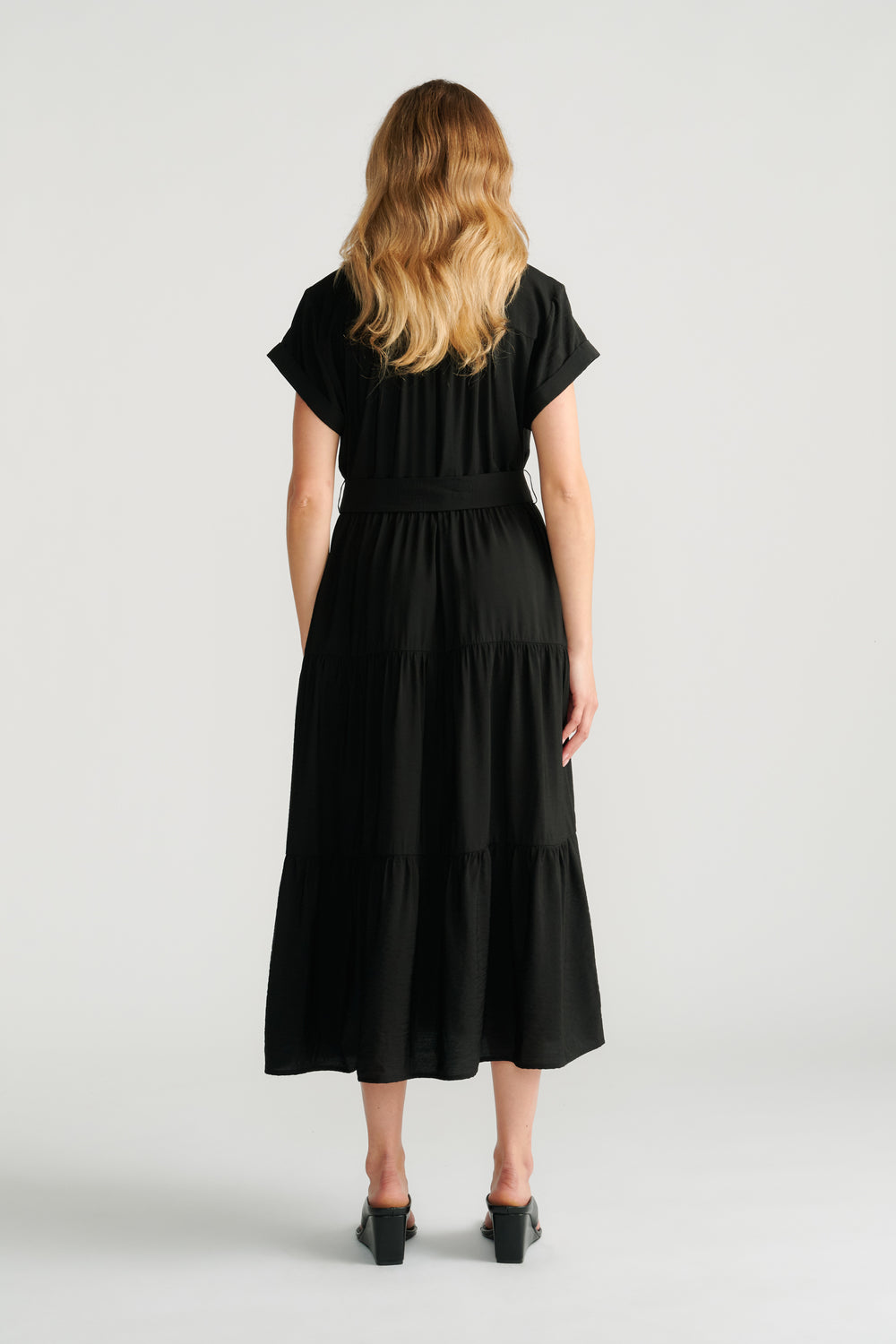 Back view of woman wearing a flowing black midi dress with short sleeves and tiered skirt, paired with block heel shoes.