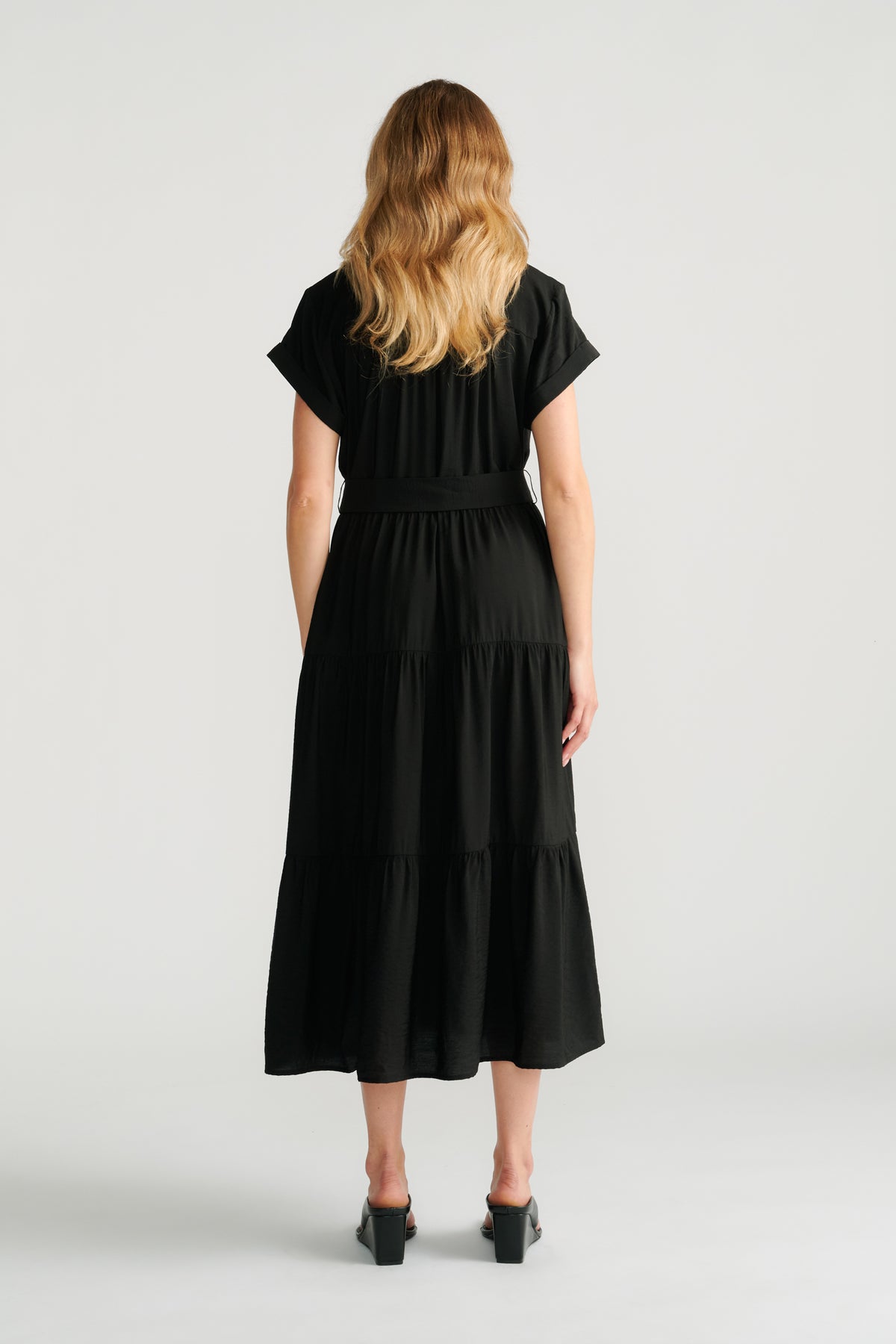 Back view of woman wearing a flowing black midi dress with short sleeves and tiered skirt, paired with block heel shoes.