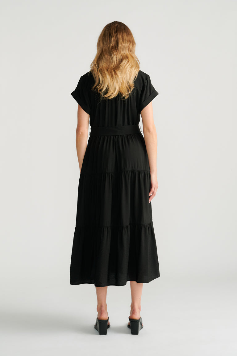 Back view of woman wearing a flowing black midi dress with short sleeves and tiered skirt, paired with block heel shoes.
