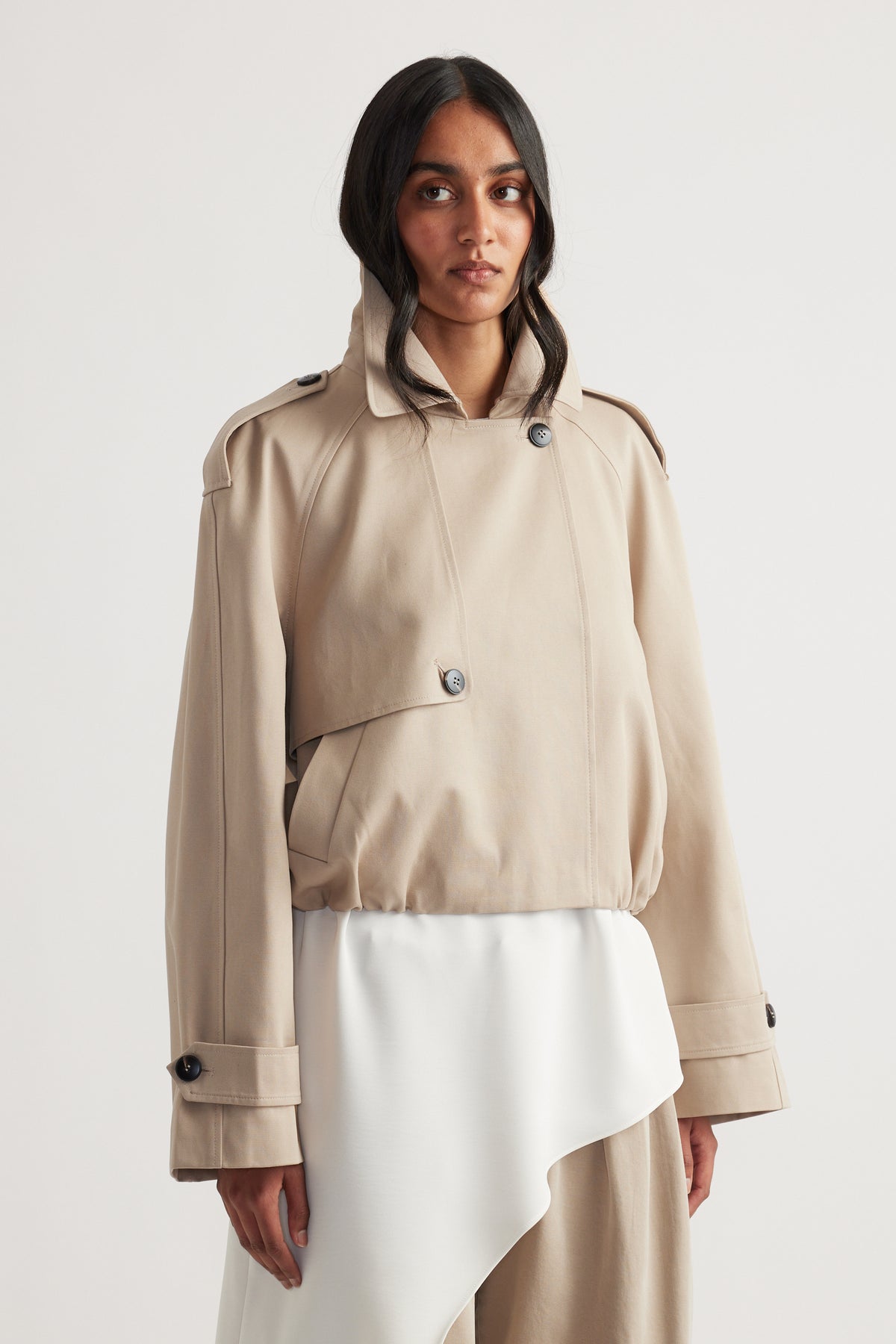 Woman wearing a light beige cropped trench coat with button details and exaggerated cuffs over a white asymmetrical dress