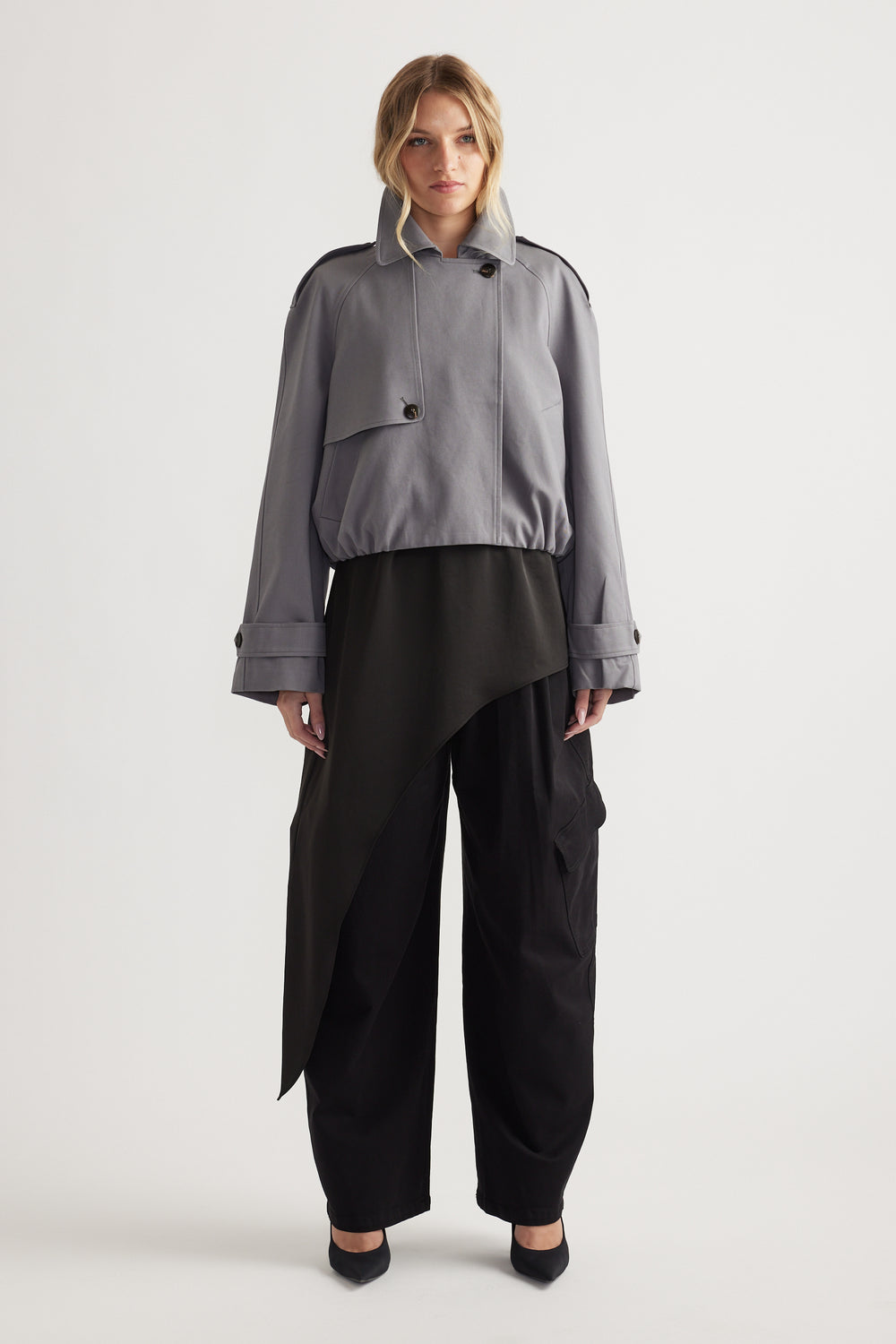 Woman wearing a grey cropped bubble trench with wide sleeves over black asymmetric layered pants and black heels