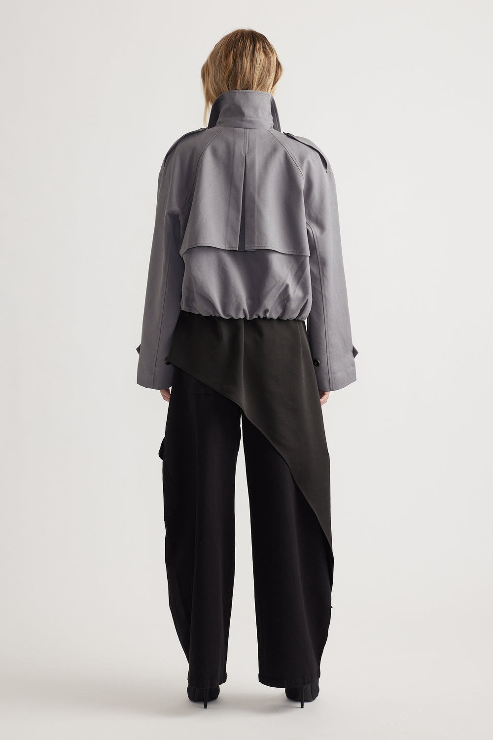 Rear view of woman wearing a grey cropped trench jacket with structured back detail and wide black pants with an asymmetrical overlay panel