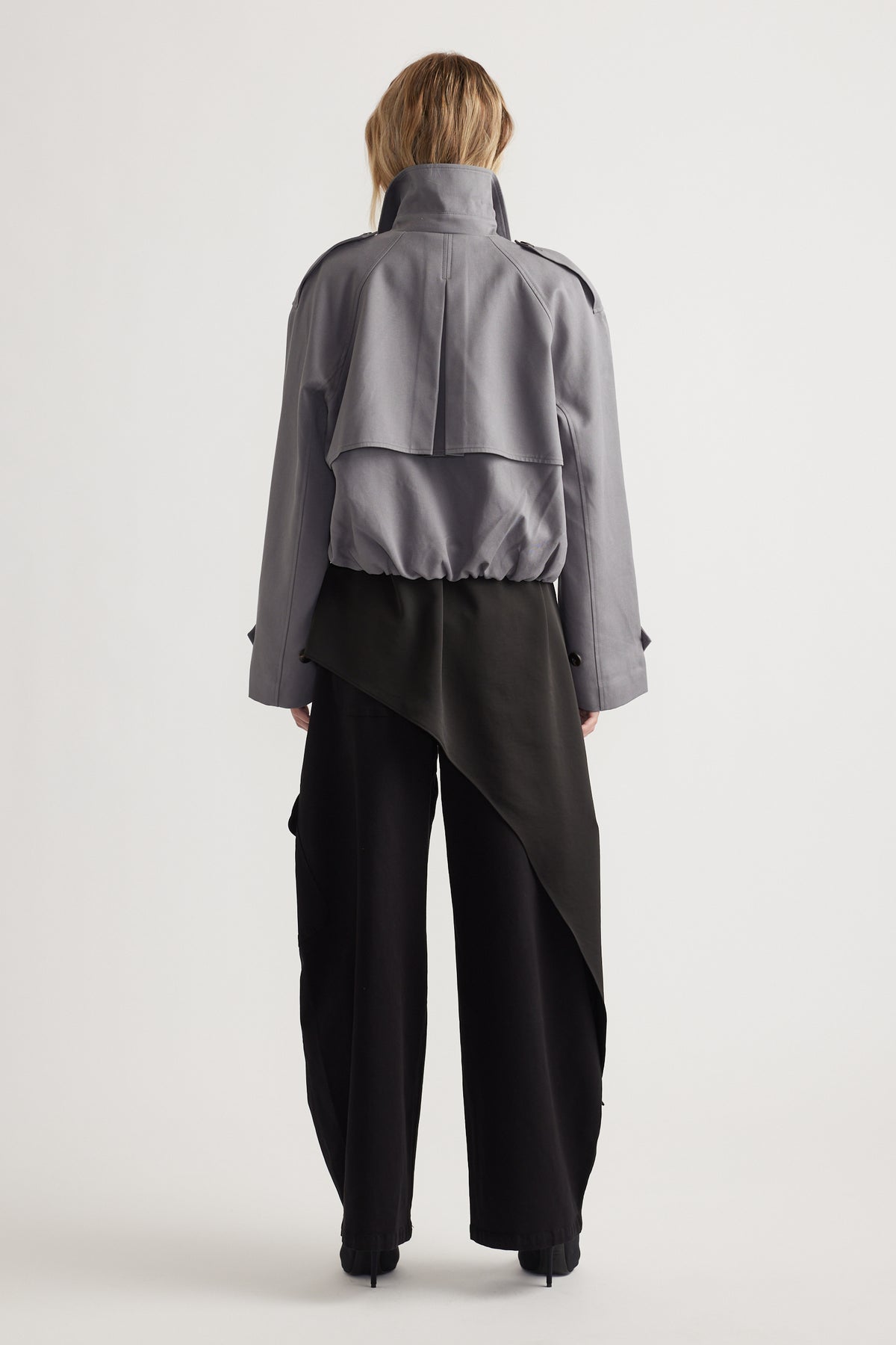Rear view of woman wearing a grey cropped trench jacket with structured back detail and wide black pants with an asymmetrical overlay panel