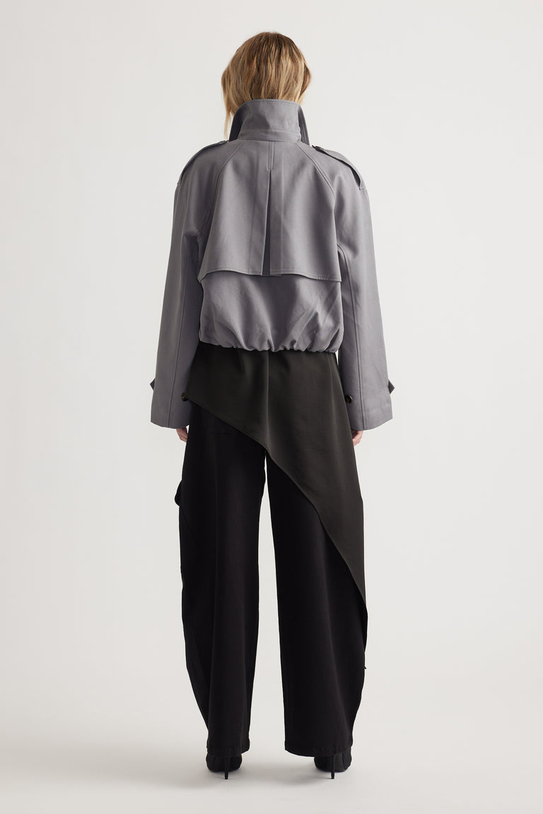 Rear view of woman wearing a grey cropped trench jacket with structured back detail and wide black pants with an asymmetrical overlay panel
