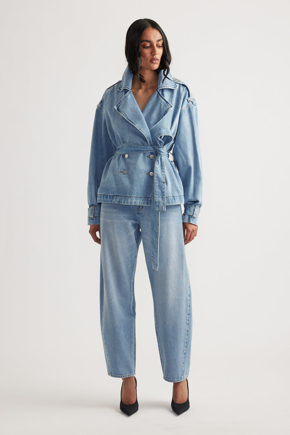 Woman wearing a belted light wash denim crop trench jacket paired with loose straight-leg jeans and black heels