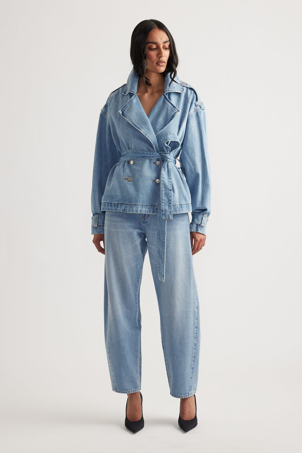 Woman wearing a belted light wash denim crop trench jacket paired with loose straight-leg jeans and black heels