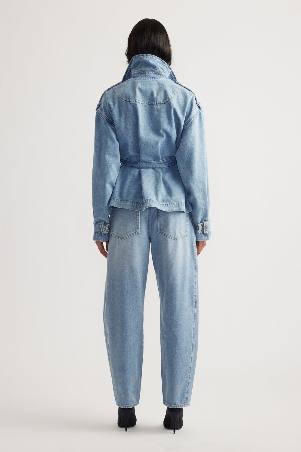 Woman seen from behind wearing a belted light wash denim jacket with a flared hem and matching high-waisted jeans