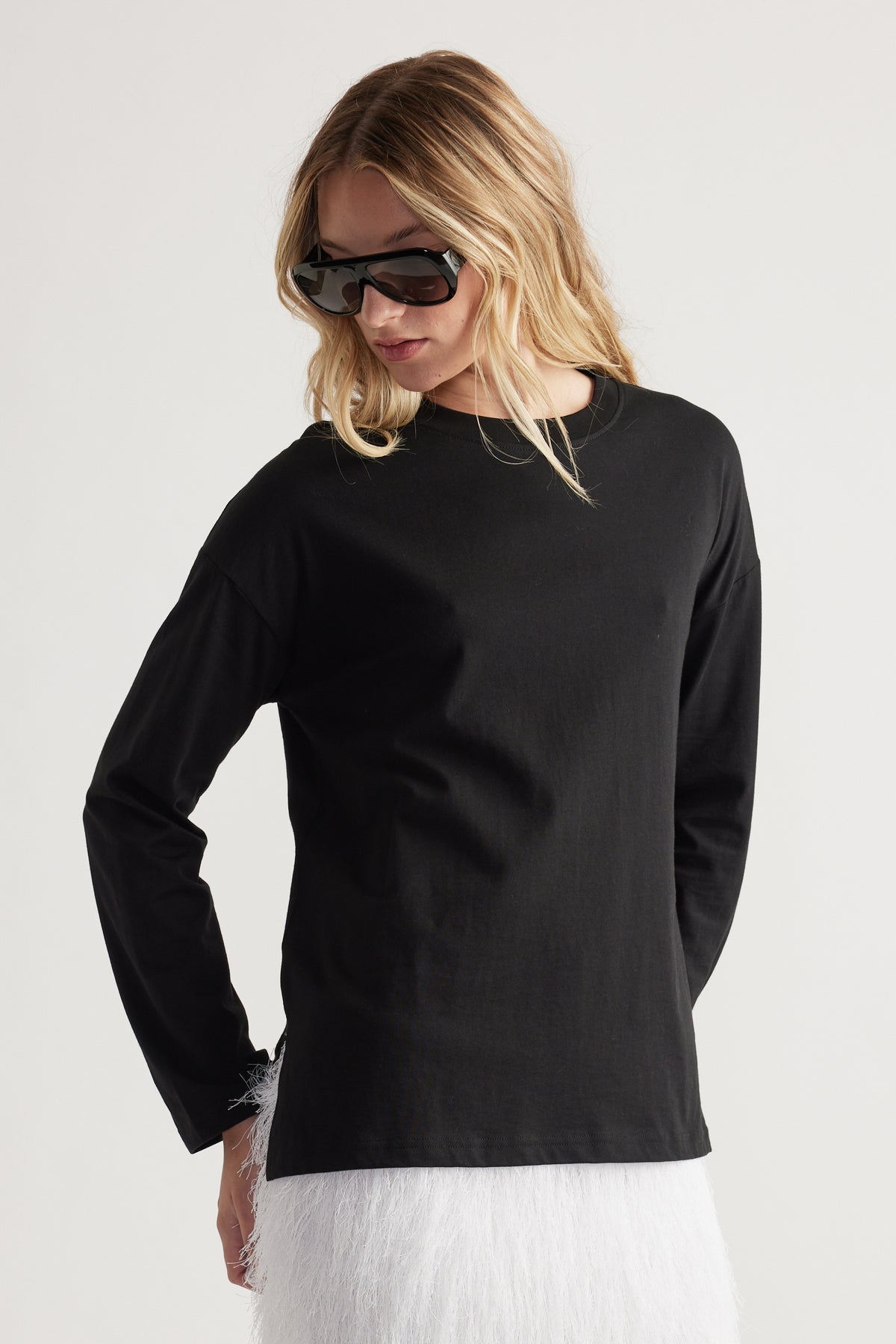Woman wearing a black long-sleeve crew neck top with a relaxed fit and oversized sunglasses, paired with a white textured skirt.