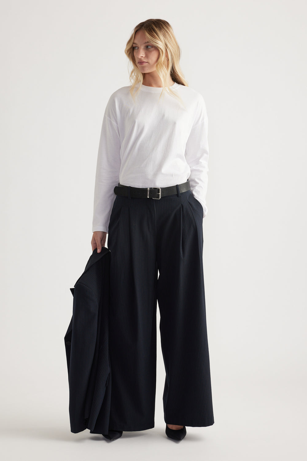 Woman wearing a white long sleeve top tucked into high-waisted wide-leg black pants, holding a matching blazer.
