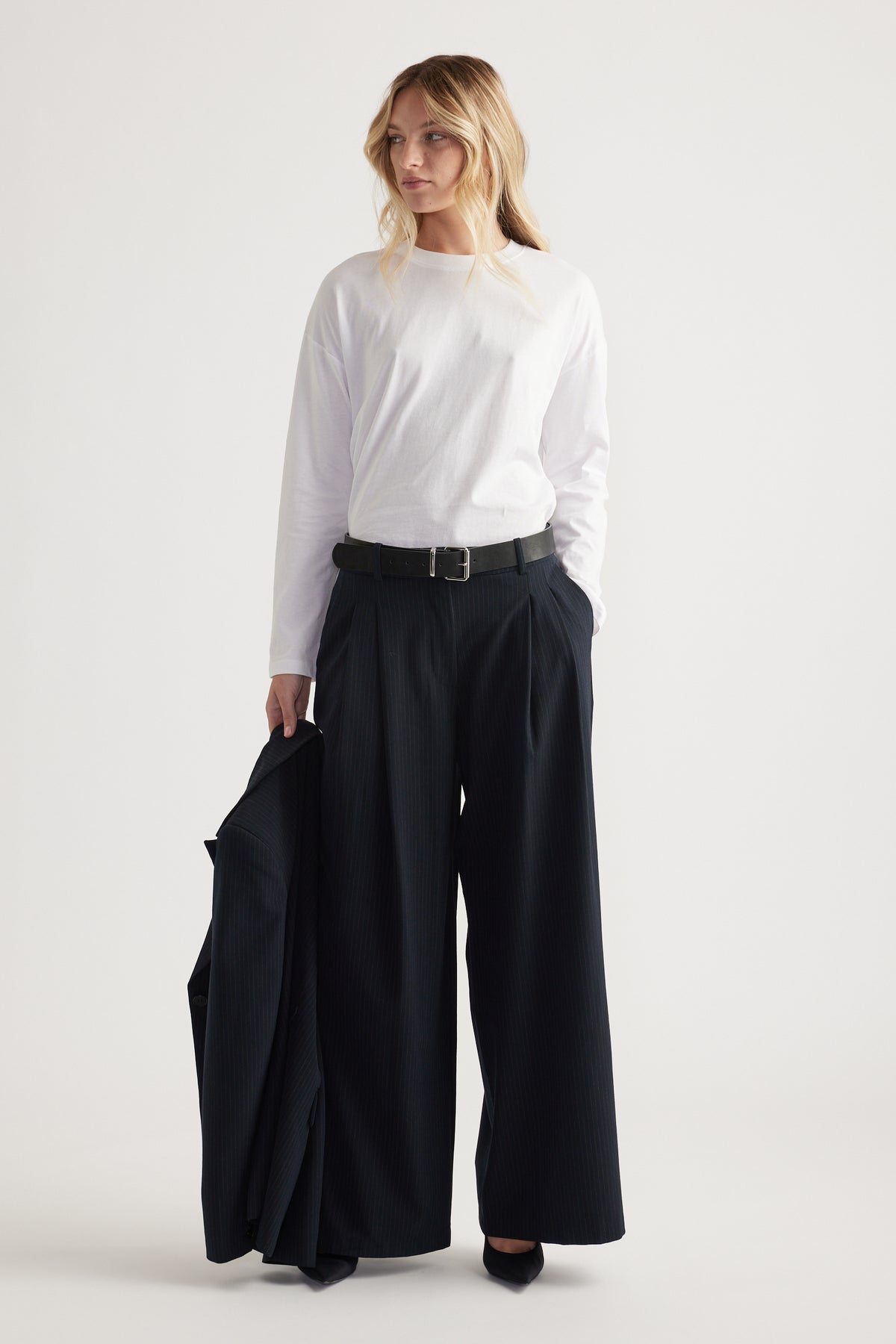 Woman wearing a white long sleeve top tucked into high-waisted wide-leg black pants, holding a matching blazer.