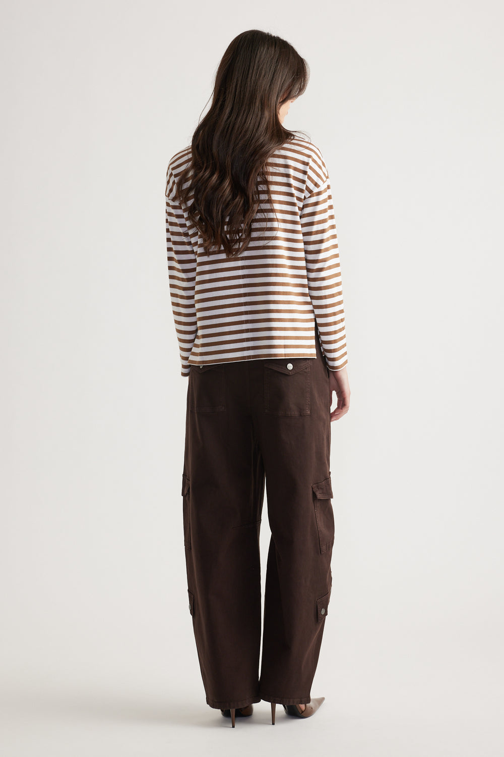 Woman wearing a tan and white striped long sleeve top paired with loose dark brown cargo pants and pointed heels, viewed from behind.