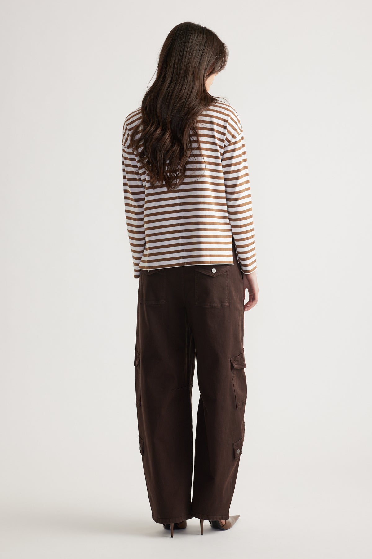 Woman wearing a tan and white striped long sleeve top paired with loose dark brown cargo pants and pointed heels, viewed from behind.