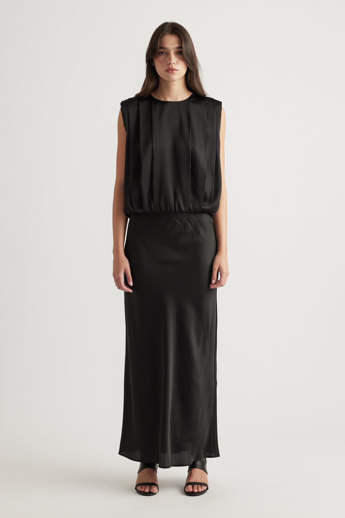 Woman wearing a sleeveless black satin dress with a relaxed top and fitted long skirt, styled with black open-toe heels