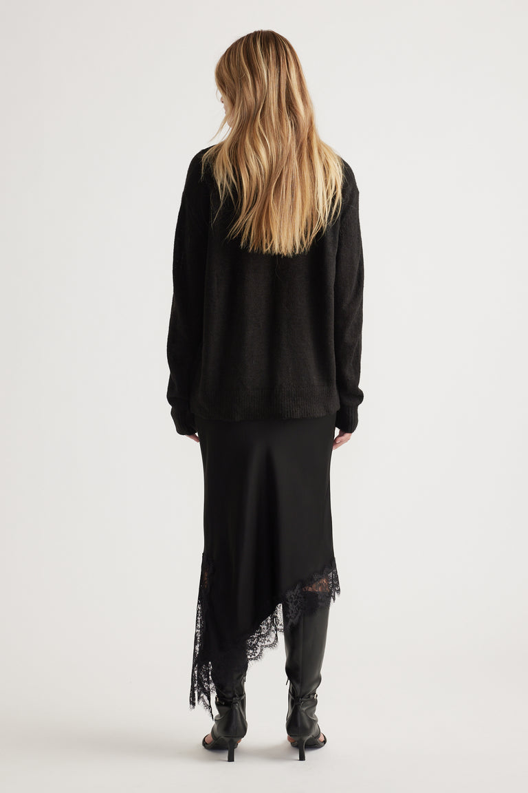 Woman wearing a relaxed black knit jumper paired with a satin asymmetrical skirt featuring lace trim and black heeled boots