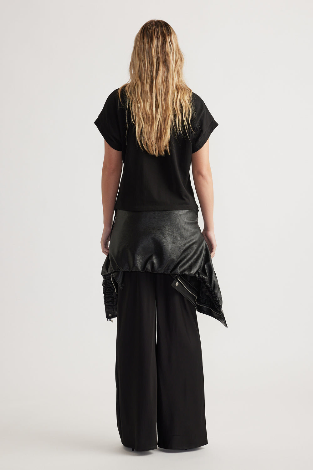 Rear view of woman wearing relaxed fit black short sleeve top with wide-leg black pants and leather jacket tied at waist