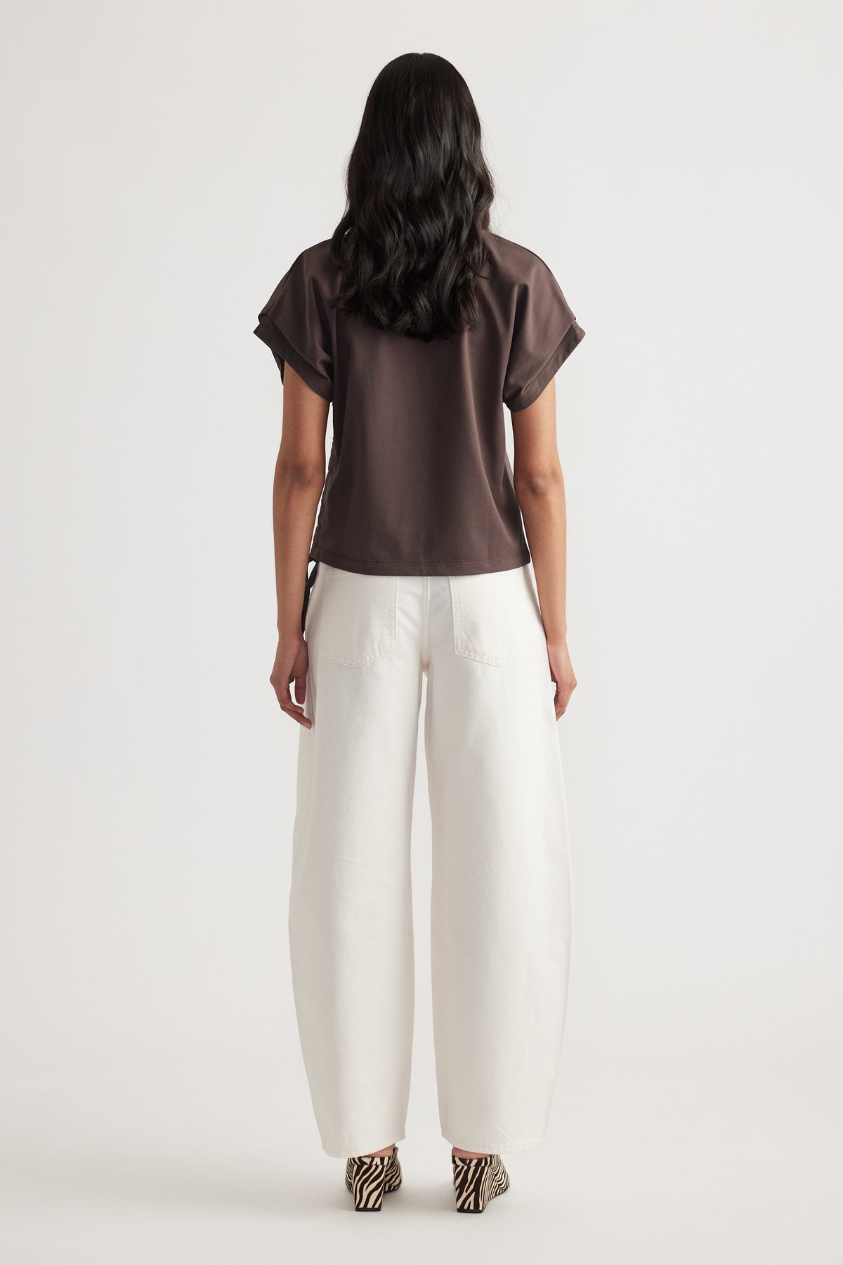 Woman wearing a relaxed fit dark top with side tie detail and wide white pants, viewed from behind on plain background