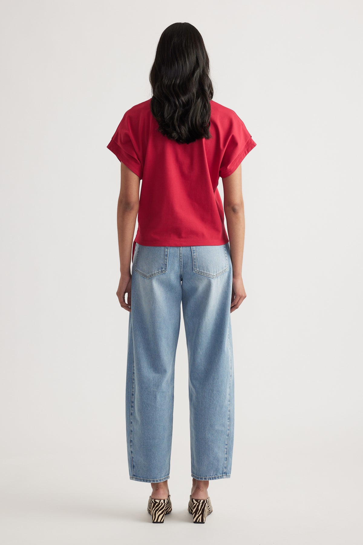 Woman wearing relaxed fit light blue jeans paired with a loose short sleeve red top, viewed from behind on plain background