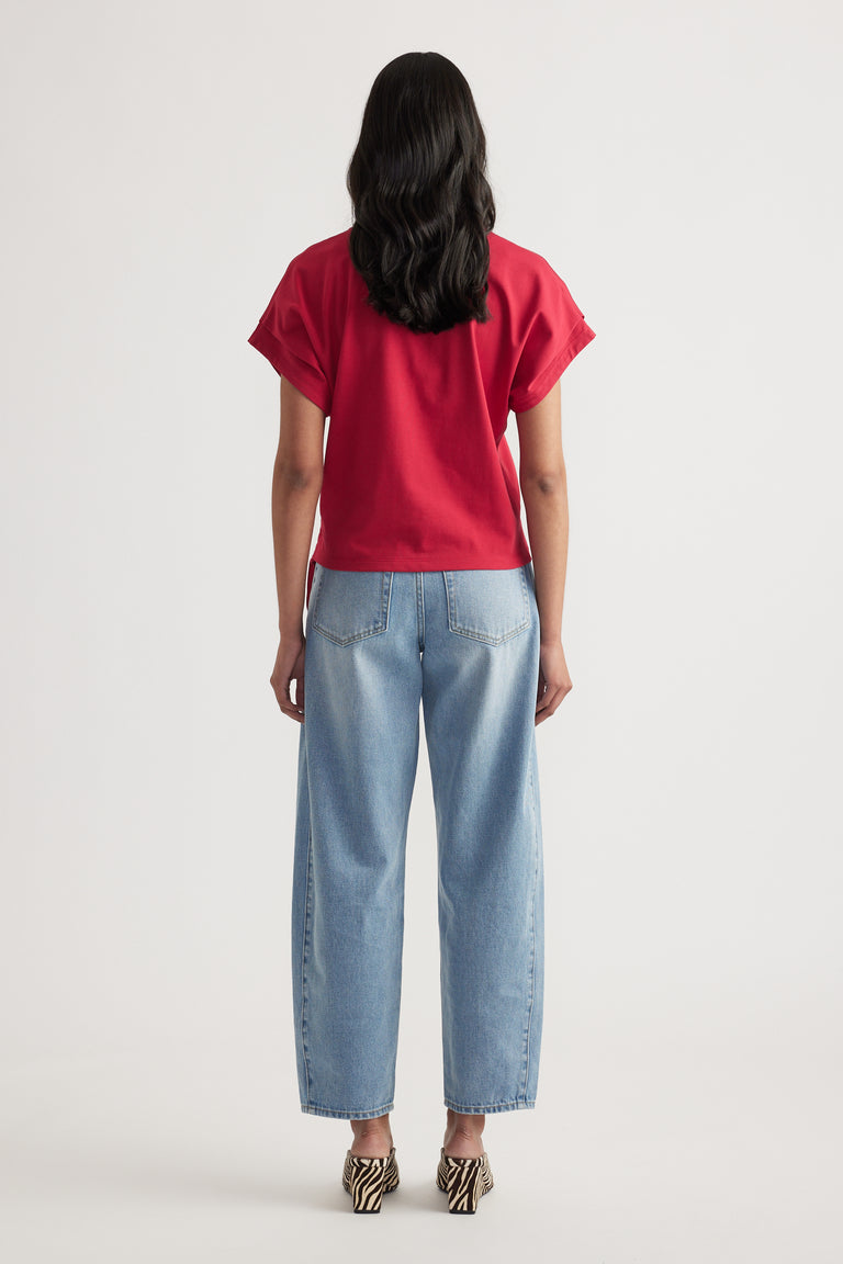 Woman wearing relaxed fit light blue jeans paired with a loose short sleeve red top, viewed from behind on plain background