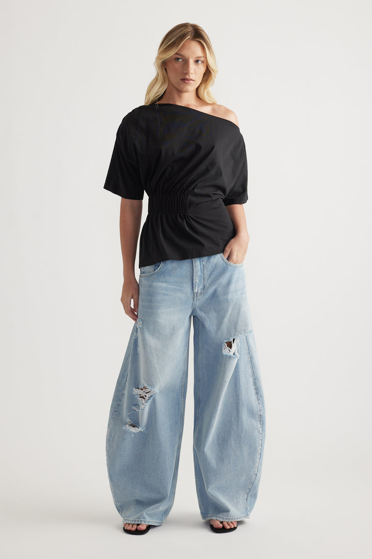 Model wearing a black asymmetrical neckline tee with a cinched waist and distressed light blue wide leg jeans