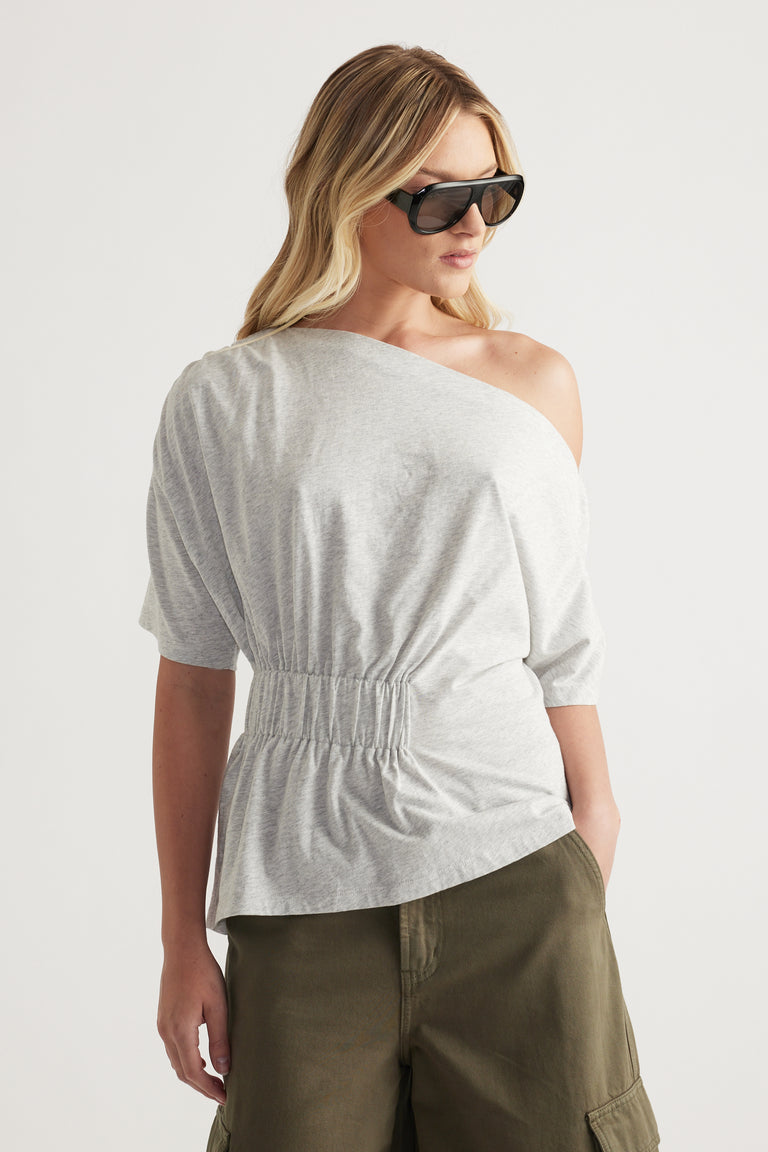 Woman wearing a light grey asymmetrical off shoulder tee with cinched waist and olive pants