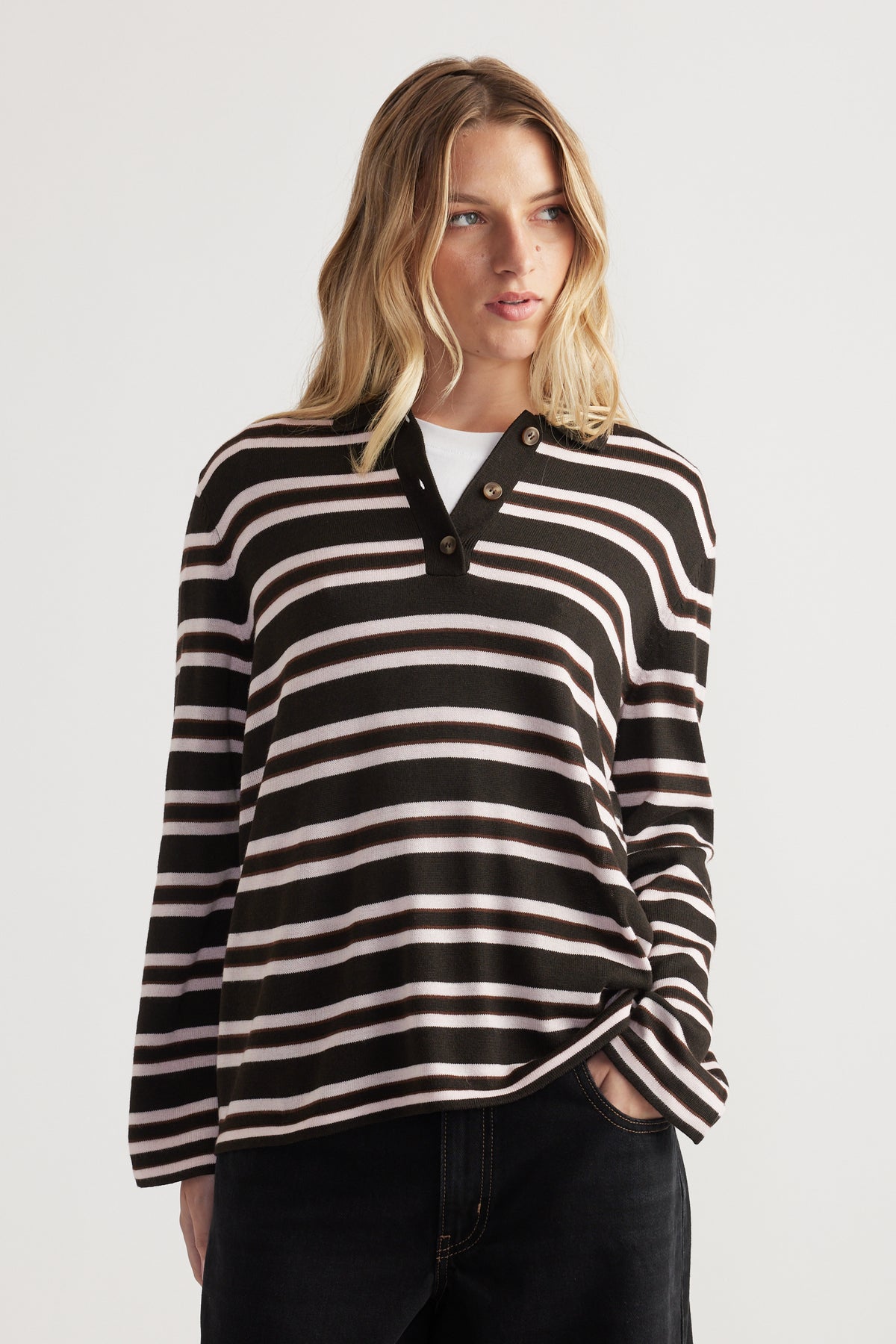 Woman wearing a striped long sleeve knit polo in black and soft pink paired with black jeans, hands in pockets
