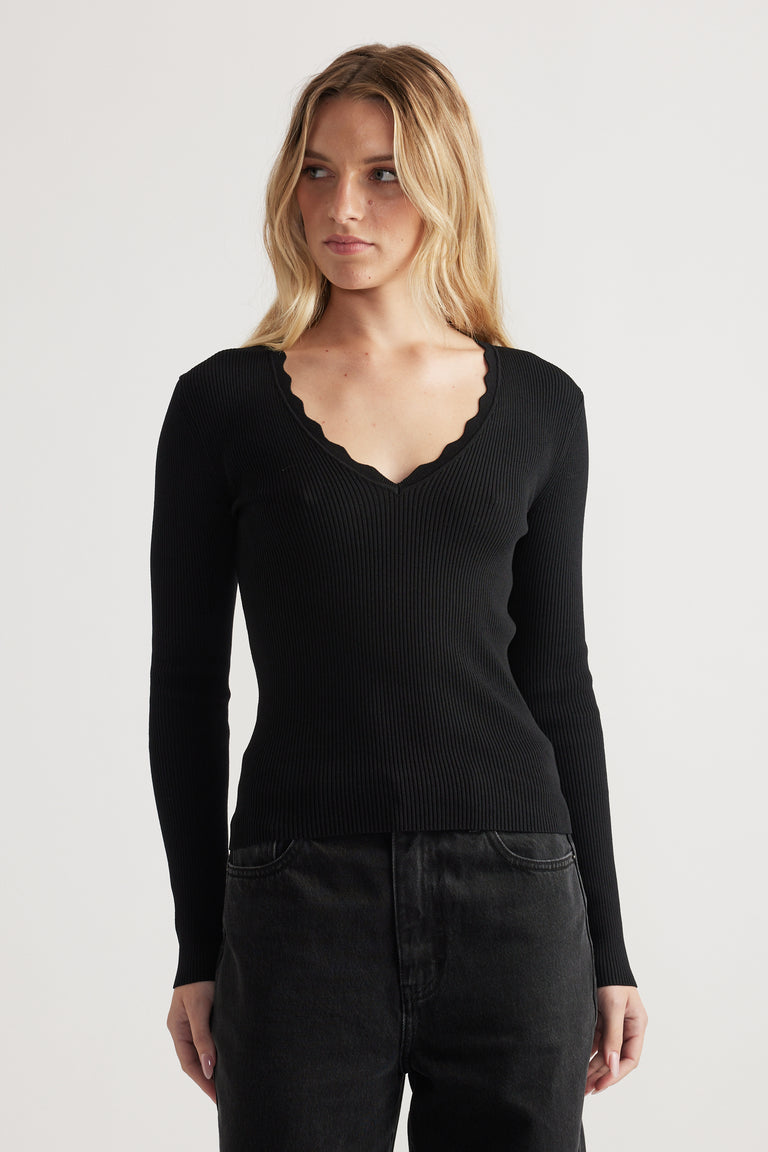 Woman wearing a fitted black ribbed long sleeve top with scalloped V neckline paired with dark jeans on plain background