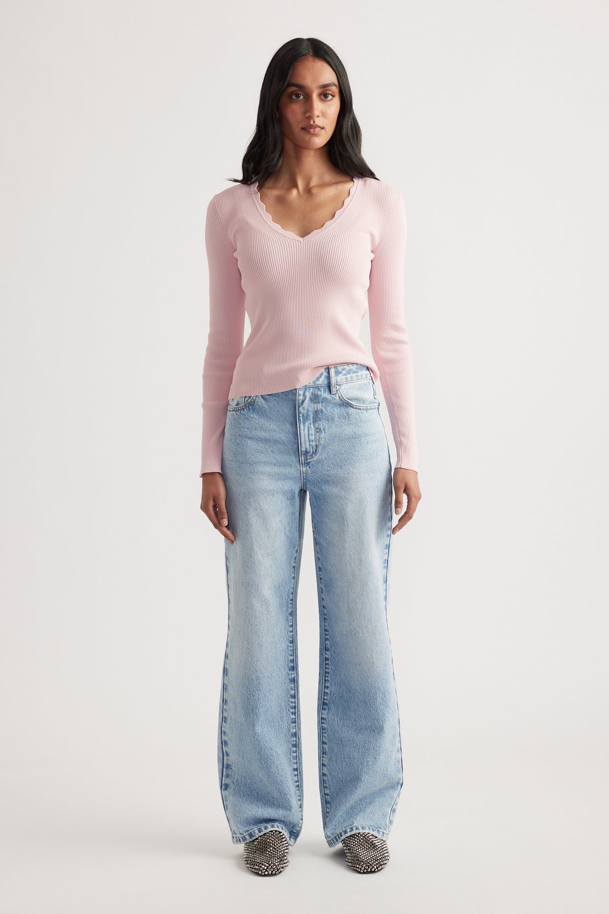 Woman wearing a fitted light pink ribbed long sleeve top with scalloped V-neck paired with wide-leg faded blue jeans