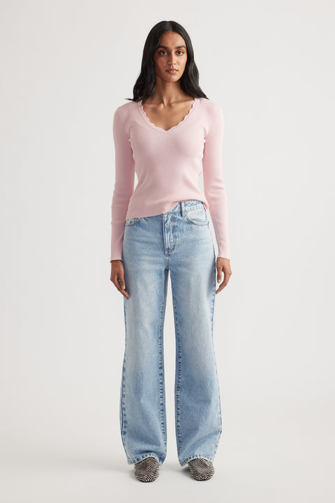 Woman wearing a fitted light pink ribbed long sleeve top with scalloped V-neck paired with wide-leg faded blue jeans