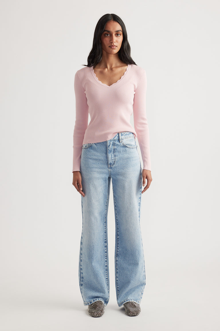 Woman wearing a fitted light pink ribbed long sleeve top with scalloped V-neck paired with wide-leg faded blue jeans