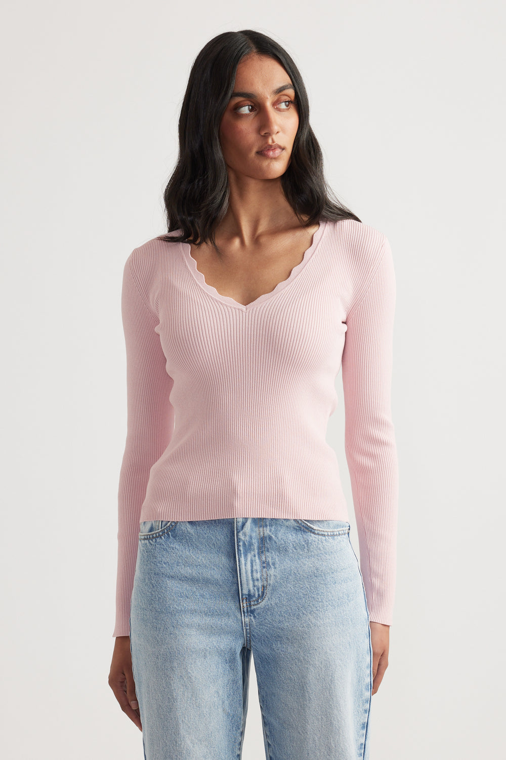 Woman wearing a fitted pale pink ribbed V-neck long sleeve top with scalloped trim paired with light wash jeans