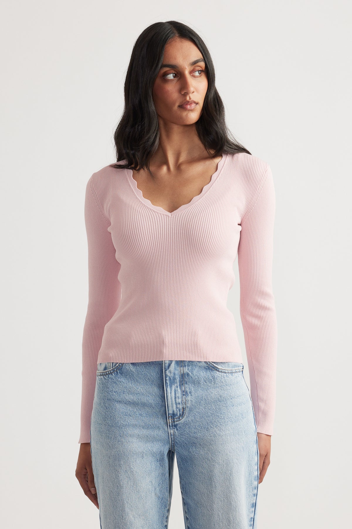 Woman wearing a fitted pale pink ribbed V-neck long sleeve top with scalloped trim paired with light wash jeans