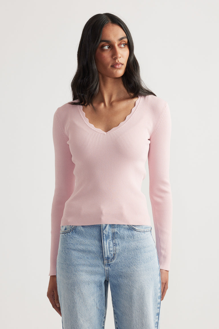 Woman wearing a fitted pale pink ribbed V-neck long sleeve top with scalloped trim paired with light wash jeans