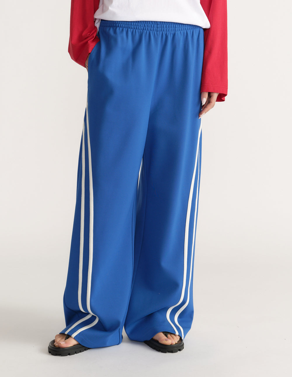 Woman wearing blue wide leg pants with white side stripes, a white tee, red long sleeve top and sandals