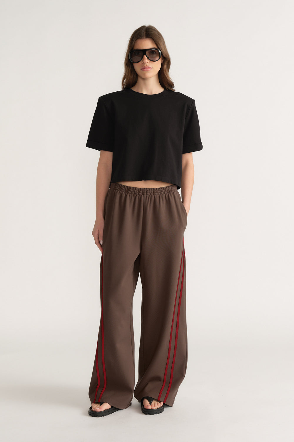 Woman in a black cropped tee and brown wide leg pants with red side stripes and sunglasses