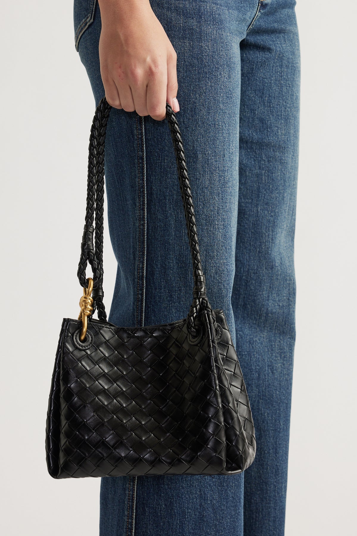 Hand holding a black woven leather bag with braided handles paired with classic blue denim jeans.