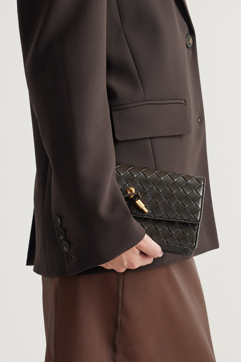 Woman holding a dark woven crossbody bag with gold hardware, paired with a tailored brown blazer and skirt.