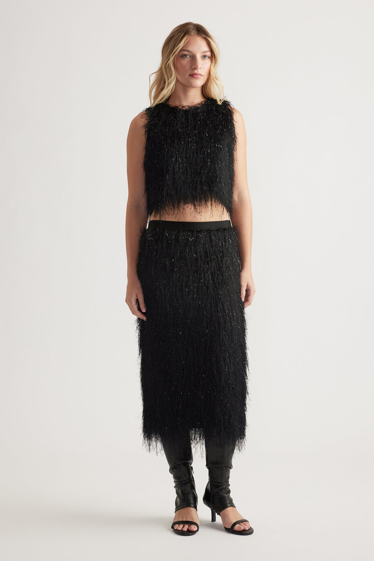 Woman wearing a black textured sleeveless top and matching midi skirt with subtle shimmer, paired with black open-toe heels
