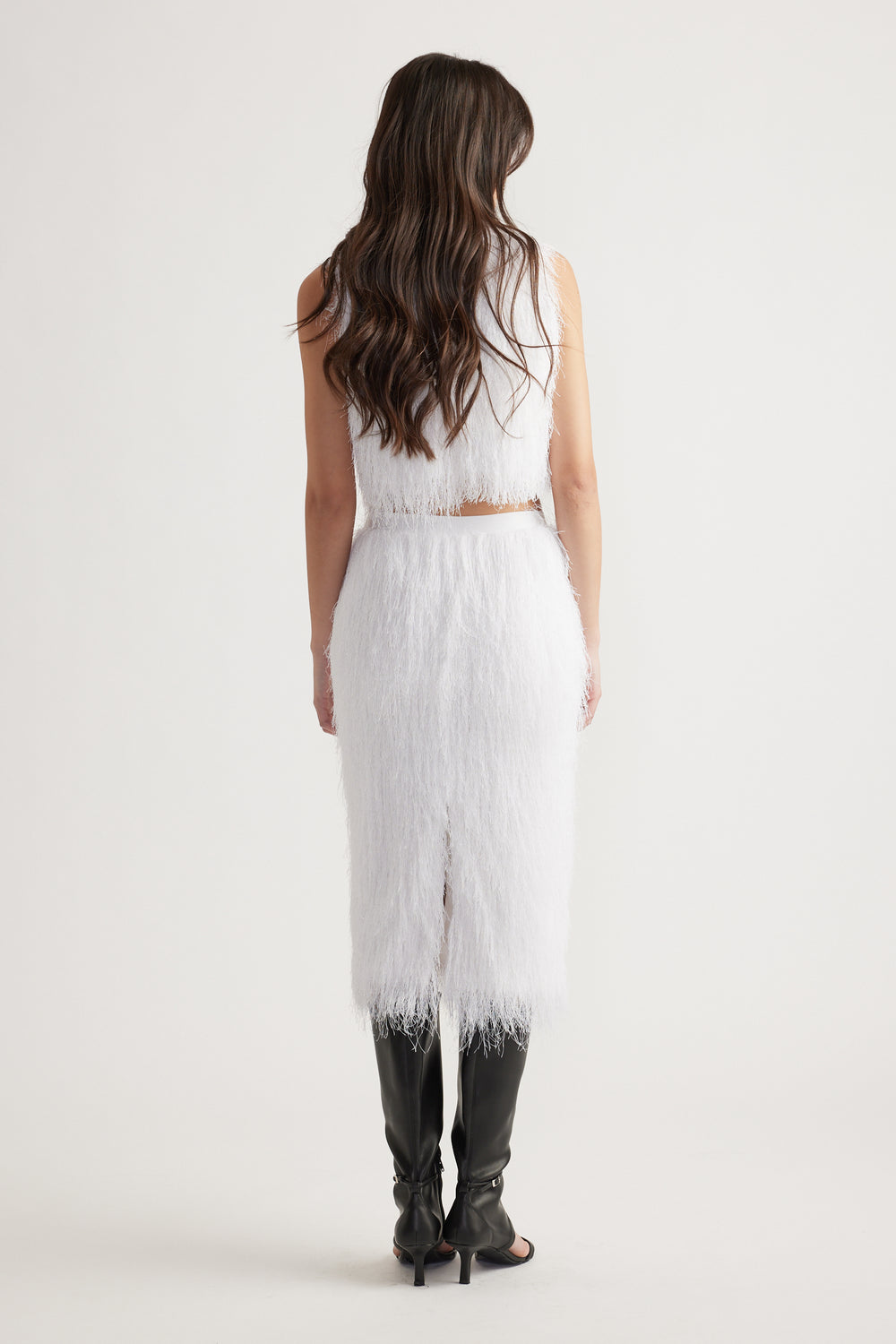 Woman wearing a textured white sleeveless top and matching midi skirt with black knee-high heeled boots, shown from behind