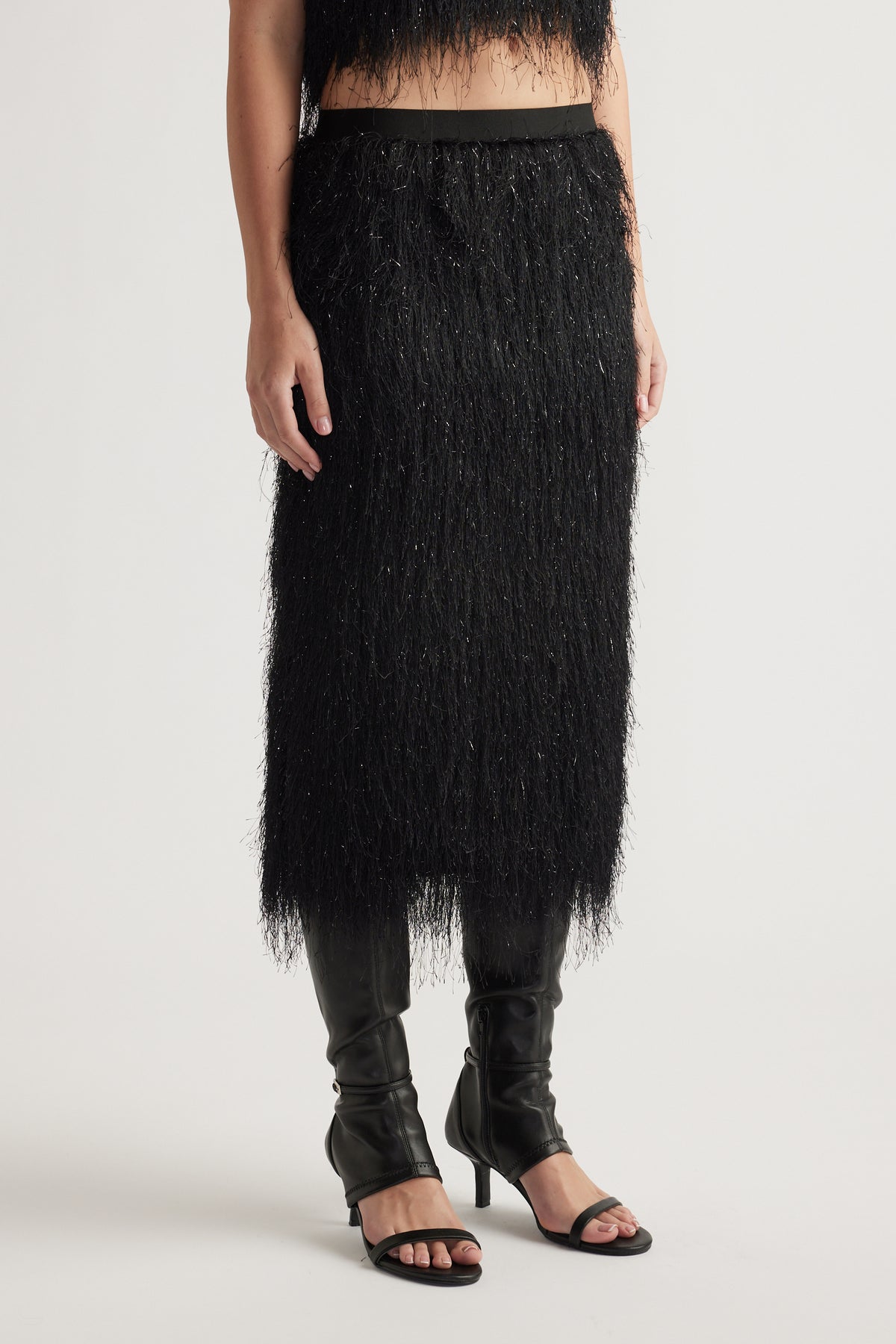 Black midi skirt with textured lurex fringe paired with open-toe heels and sleek knee-high boots.