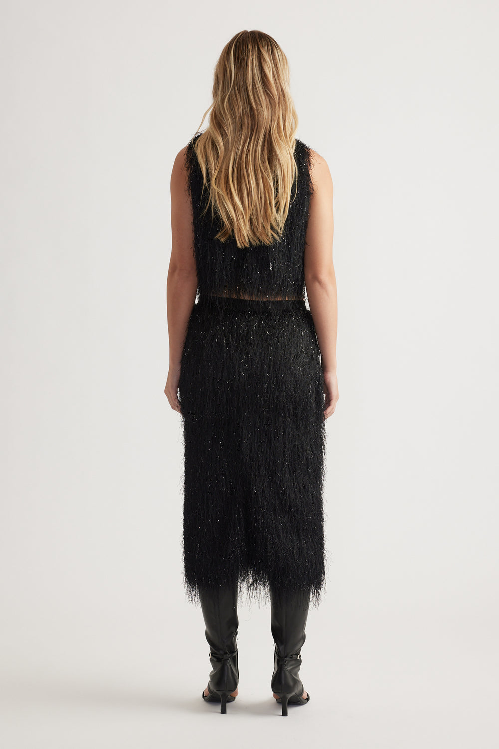 Woman seen from behind wearing a black fringed midi skirt and matching sleeveless top with black heeled boots