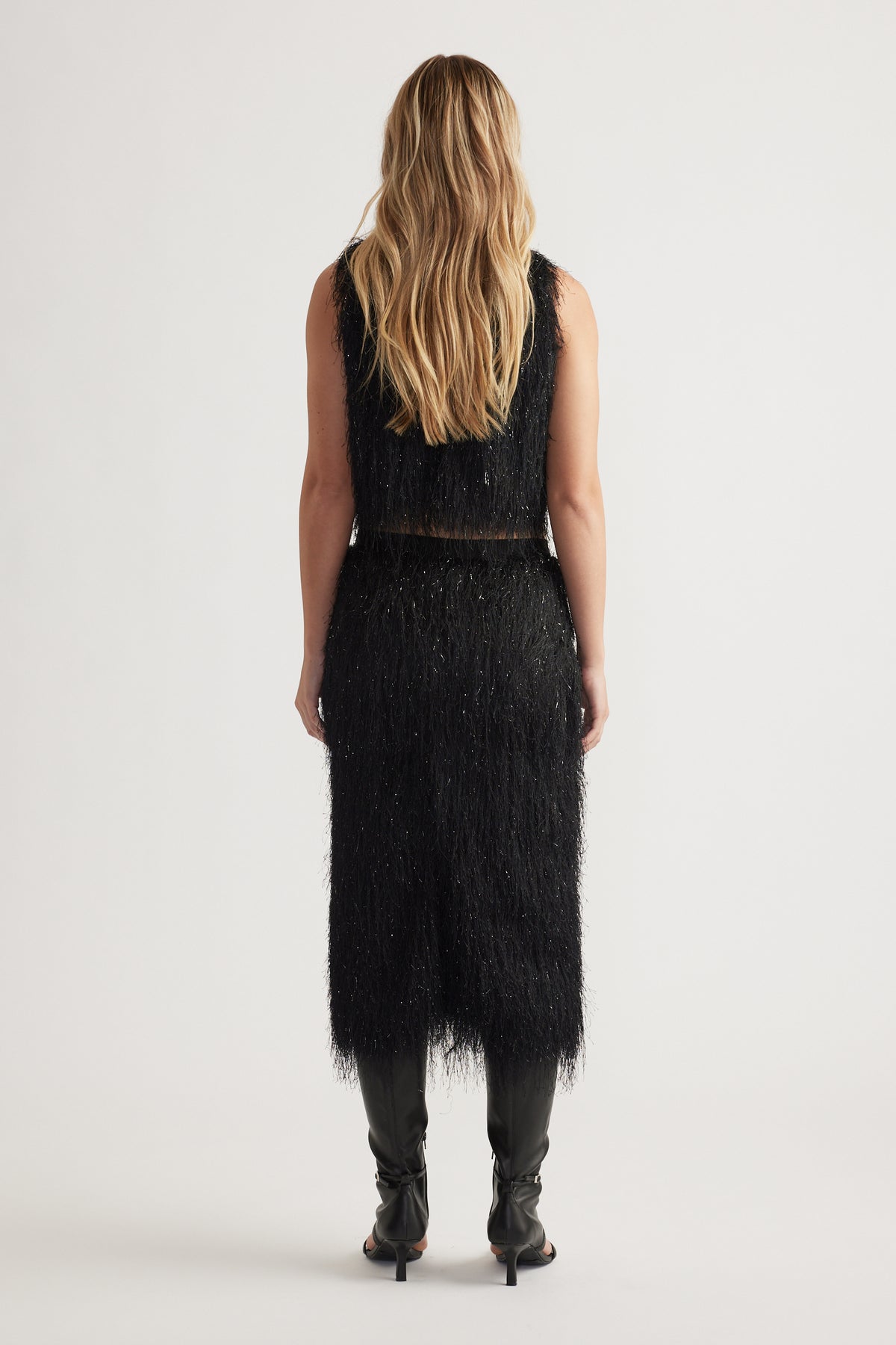Woman seen from behind wearing a black fringed midi skirt and matching sleeveless top with black heeled boots