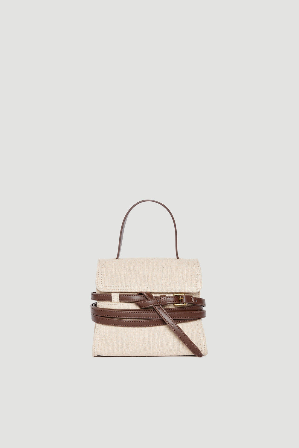 Small beige fabric bag with dark brown leather buckle, top handle, and adjustable crossbody strap on plain background