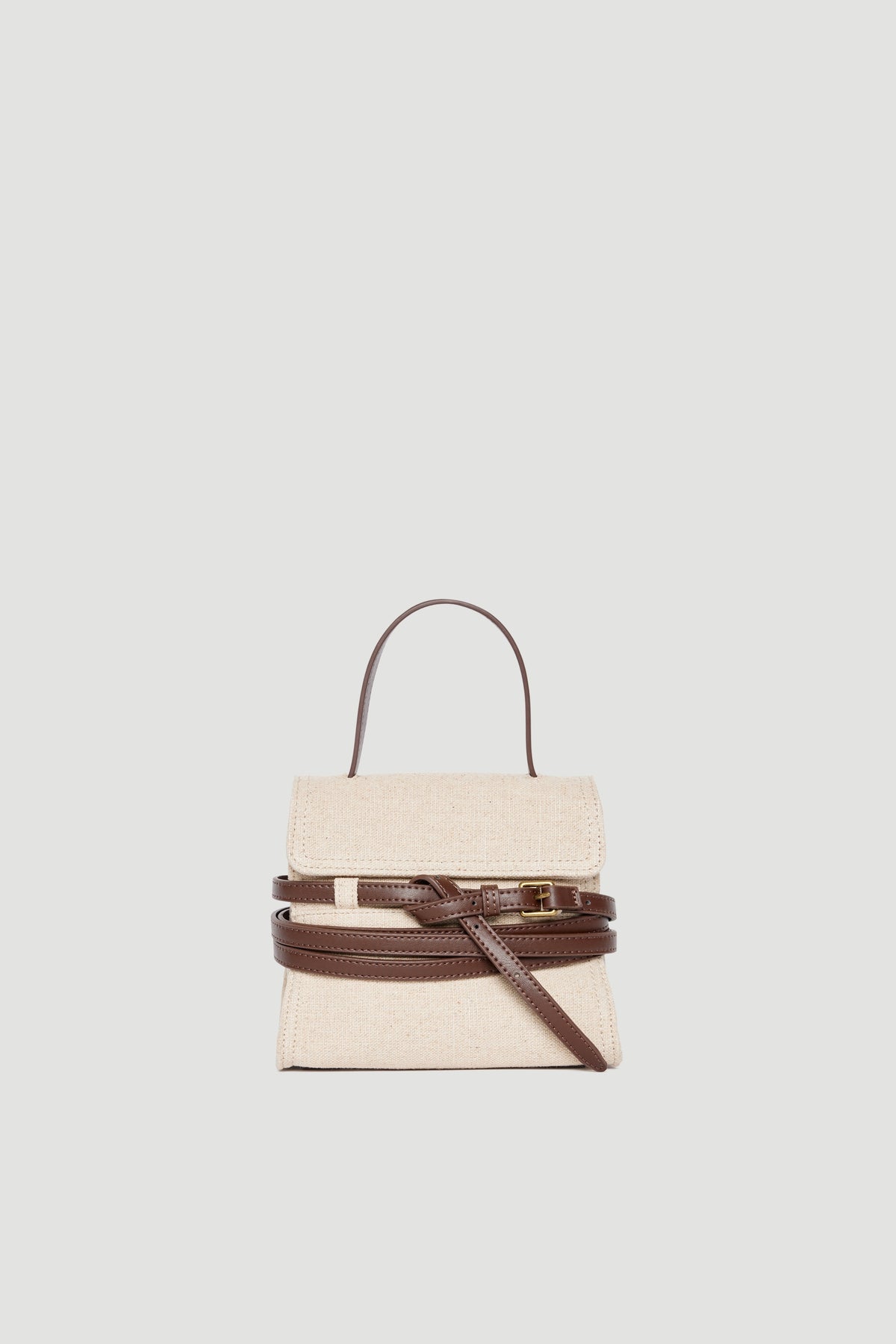 Small beige fabric bag with dark brown leather buckle, top handle, and adjustable crossbody strap on plain background