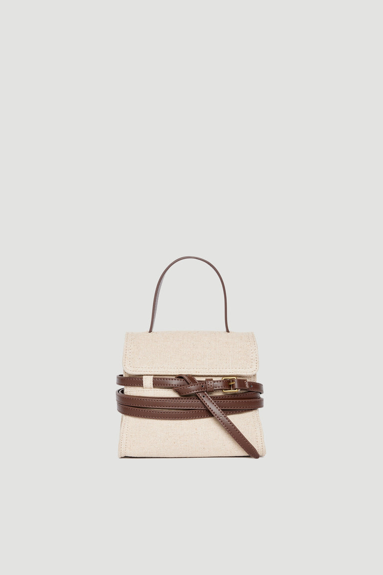 Small beige fabric bag with dark brown leather buckle, top handle, and adjustable crossbody strap on plain background