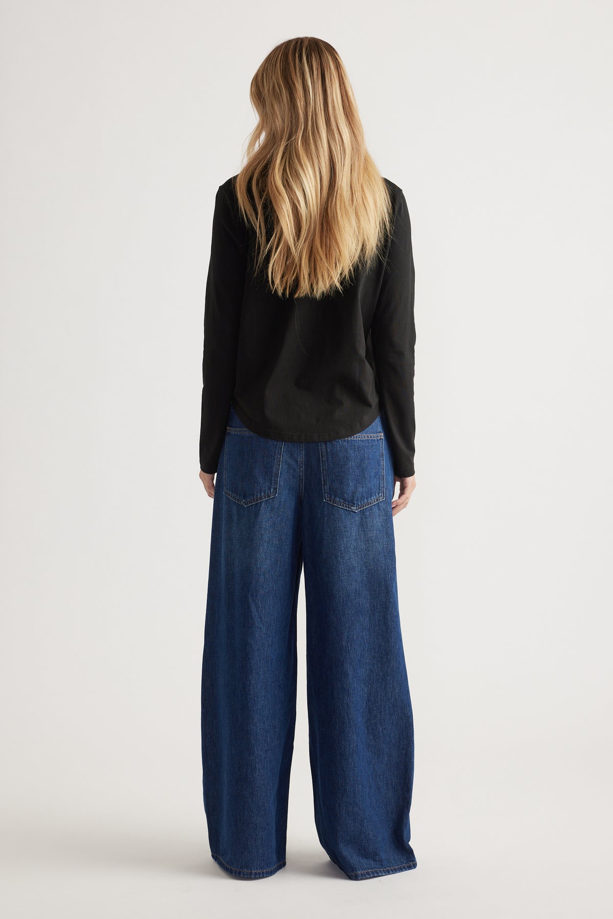 Woman with long blonde hair wearing a black long sleeve top and wide-leg dark blue denim jeans, shown from behind.