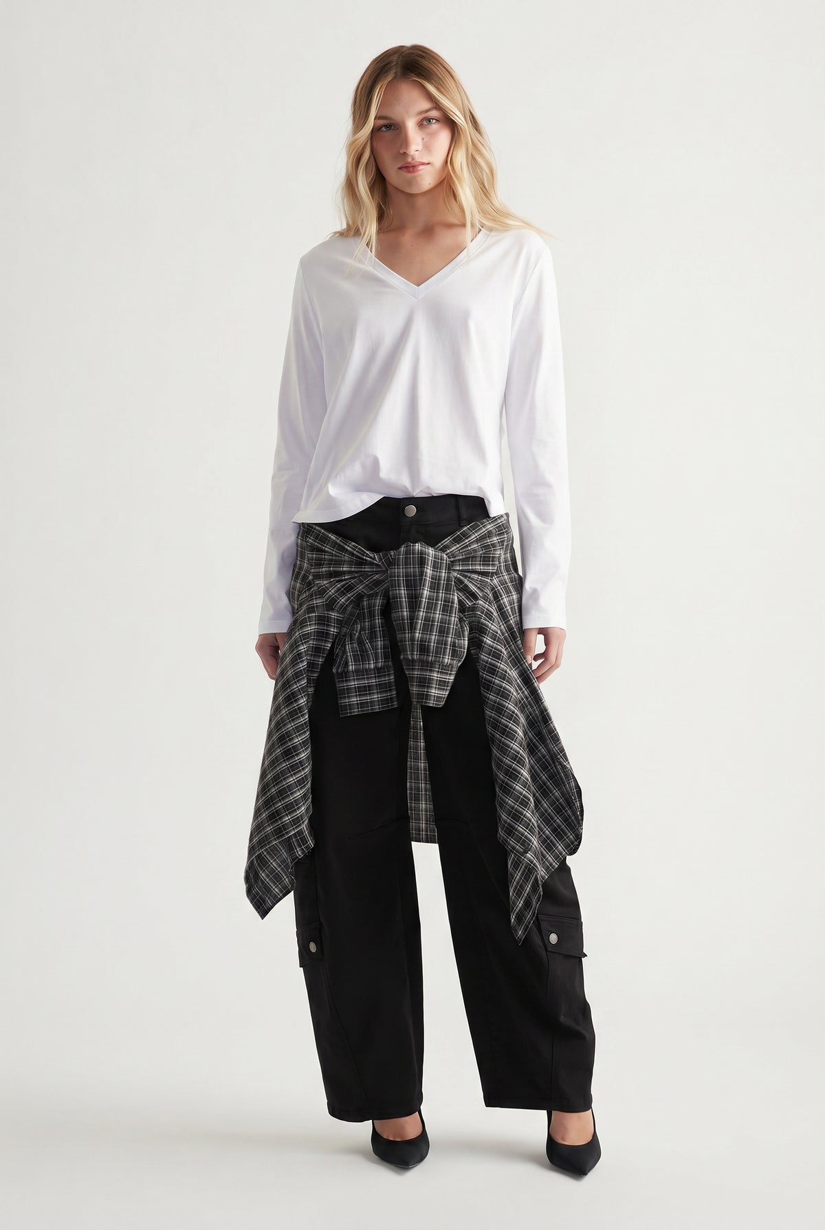 Woman wearing a white V-neck long sleeve top paired with black cargo pants and a tied plaid shirt at the waist.